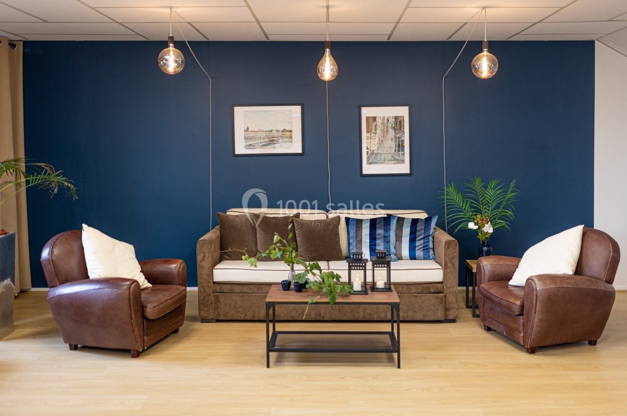 Salon avec canapé beige, fauteuils en cuir marron, table basse en bois, mur bleu décoré de cadres et plantes vertes.