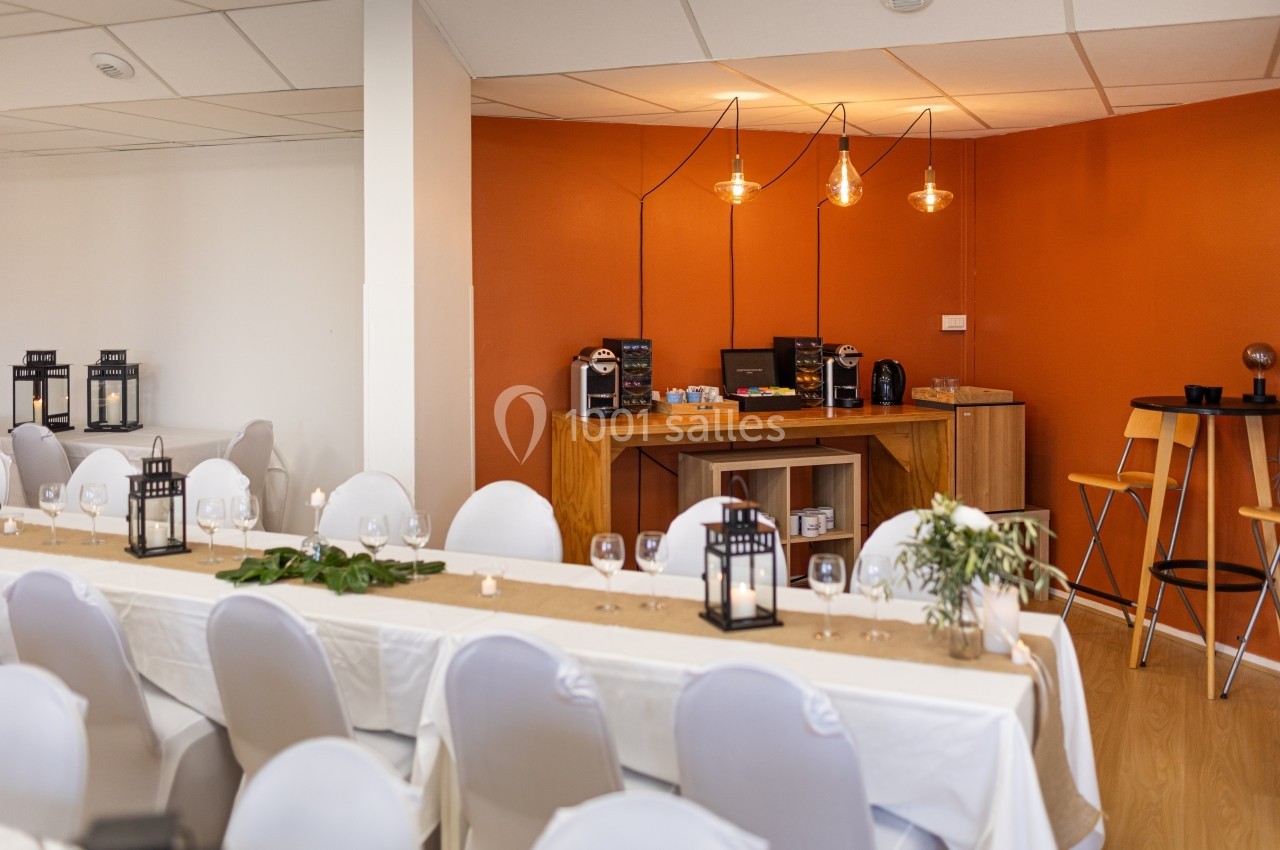 Salle décorée pour un événement, avec tables dressées, chaises blanches et un coin buffet éclairé par des suspensions.