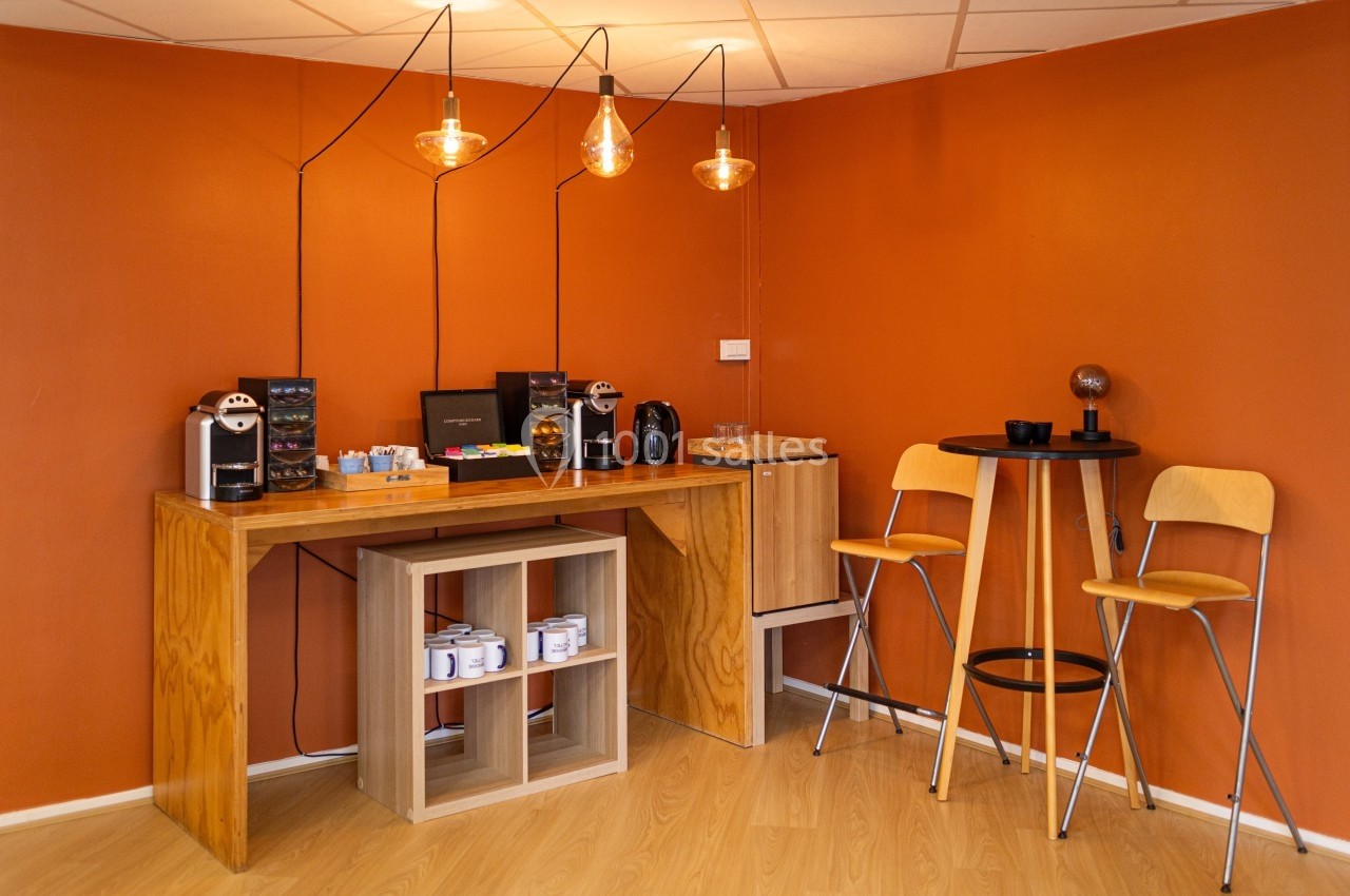 Coin café avec table en bois, machines à café, étagères avec tasses et tabourets hauts dans une pièce aux murs orange.
