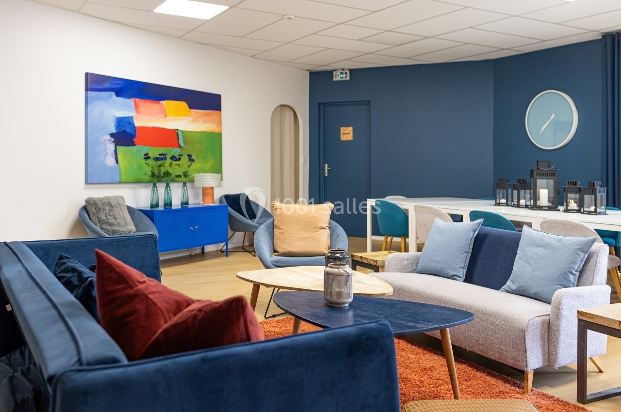 Espace salon moderne avec canapés colorés, tables basses en bois, tableau abstrait et mur bleu en arrière-plan.