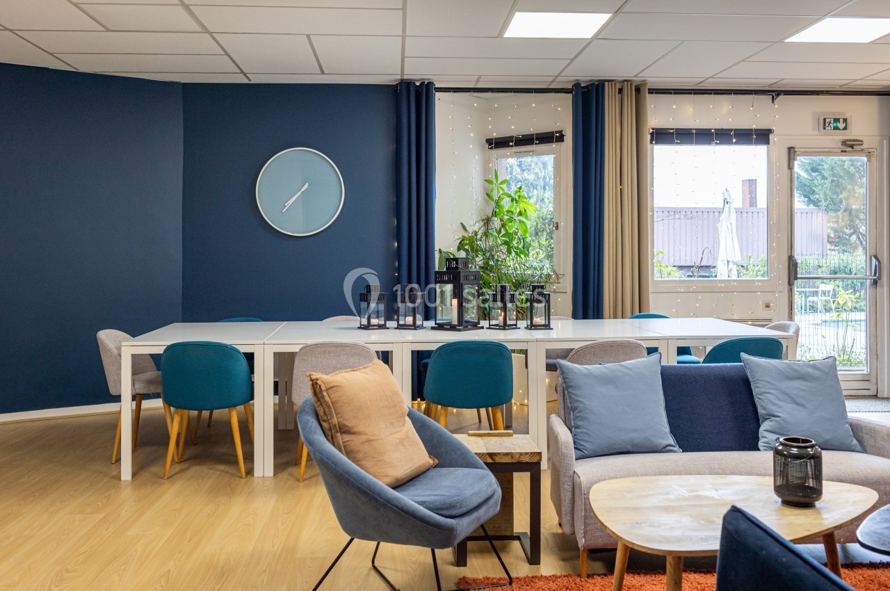 Salle lumineuse avec tables blanches, chaises colorées, canapé, plantes et grande horloge murale bleue.
