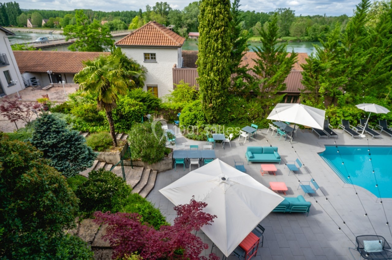 Vue d'une terrasse avec piscine, parasols, mobilier de jardin et végétation luxuriante entourée de bâtiments.