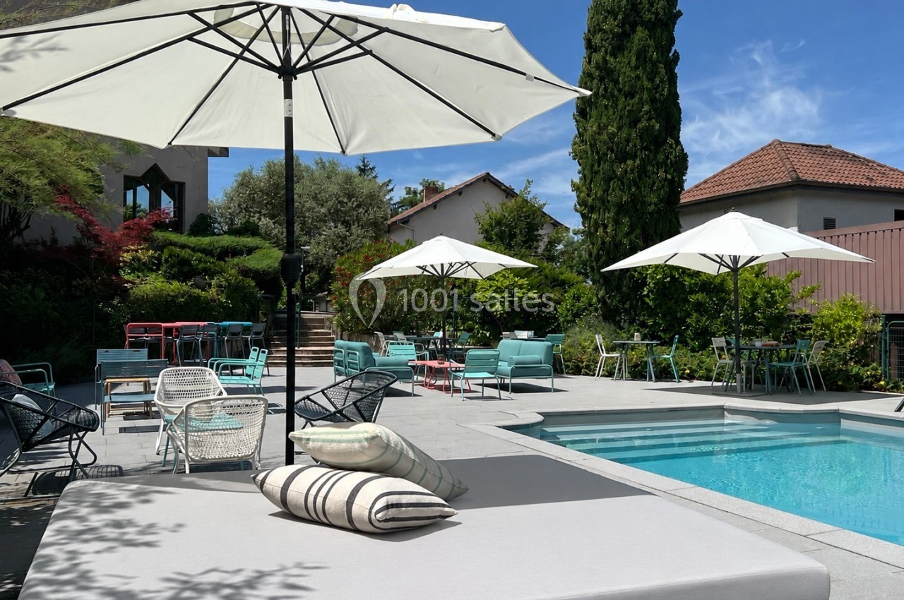 Terrasse ensoleillée avec piscine, transats, parasols blancs et mobilier de jardin entourés de verdure et maisons.