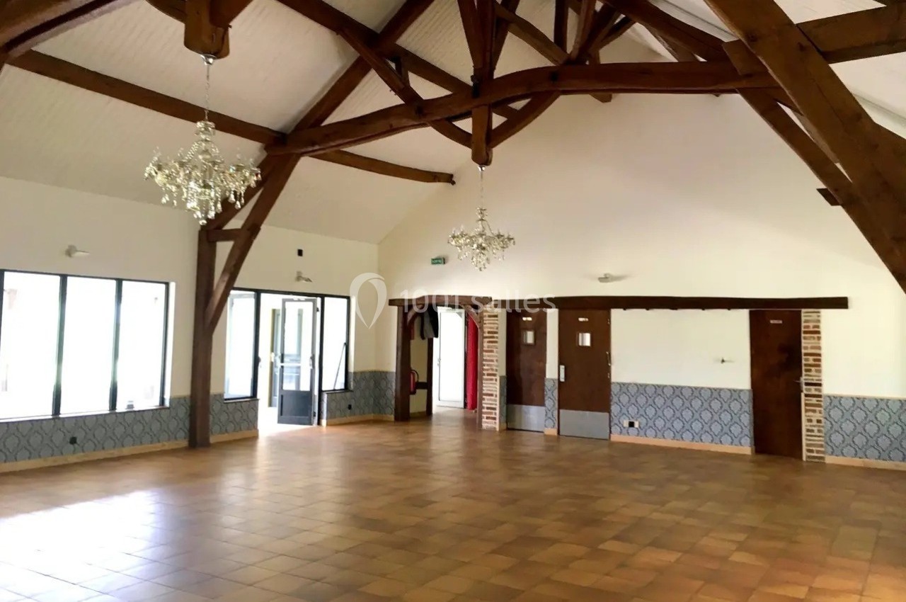 Salle vide avec poutres apparentes, carrelage au sol, murs clairs, lustres suspendus et grandes ouvertures vitrées.