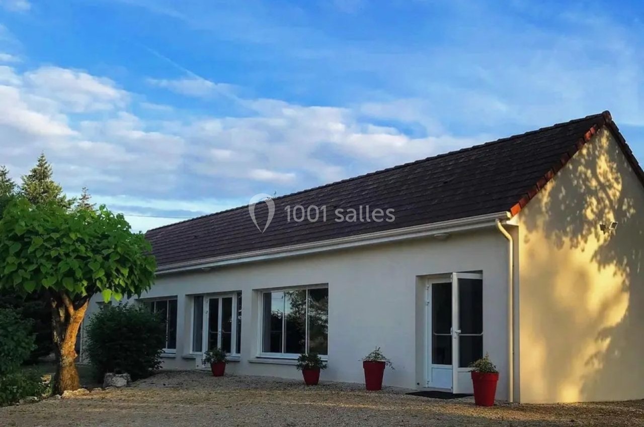 Maison de plain-pied avec façade blanche, toit en tuiles brunes, entourée de végétation et éclairée par le soleil.