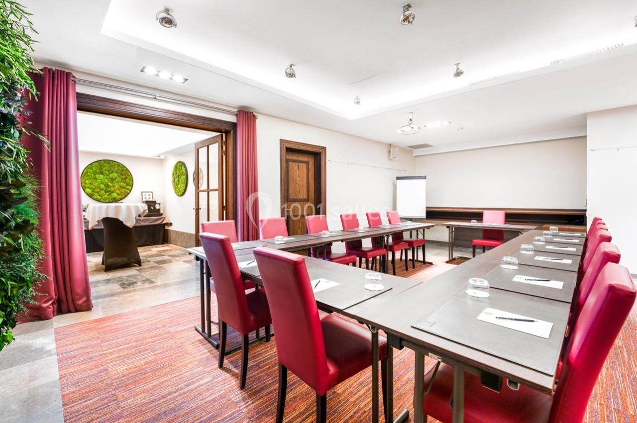 Salle de réunion lumineuse avec tables rectangulaires, chaises rouges, tableau blanc et décoration végétale murale.