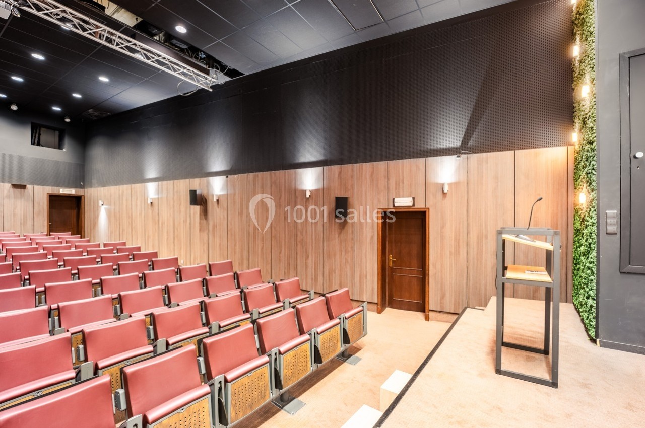Salle de conférence avec rangées de sièges rouges, pupitre en bois et mur décoré de panneaux et végétation.