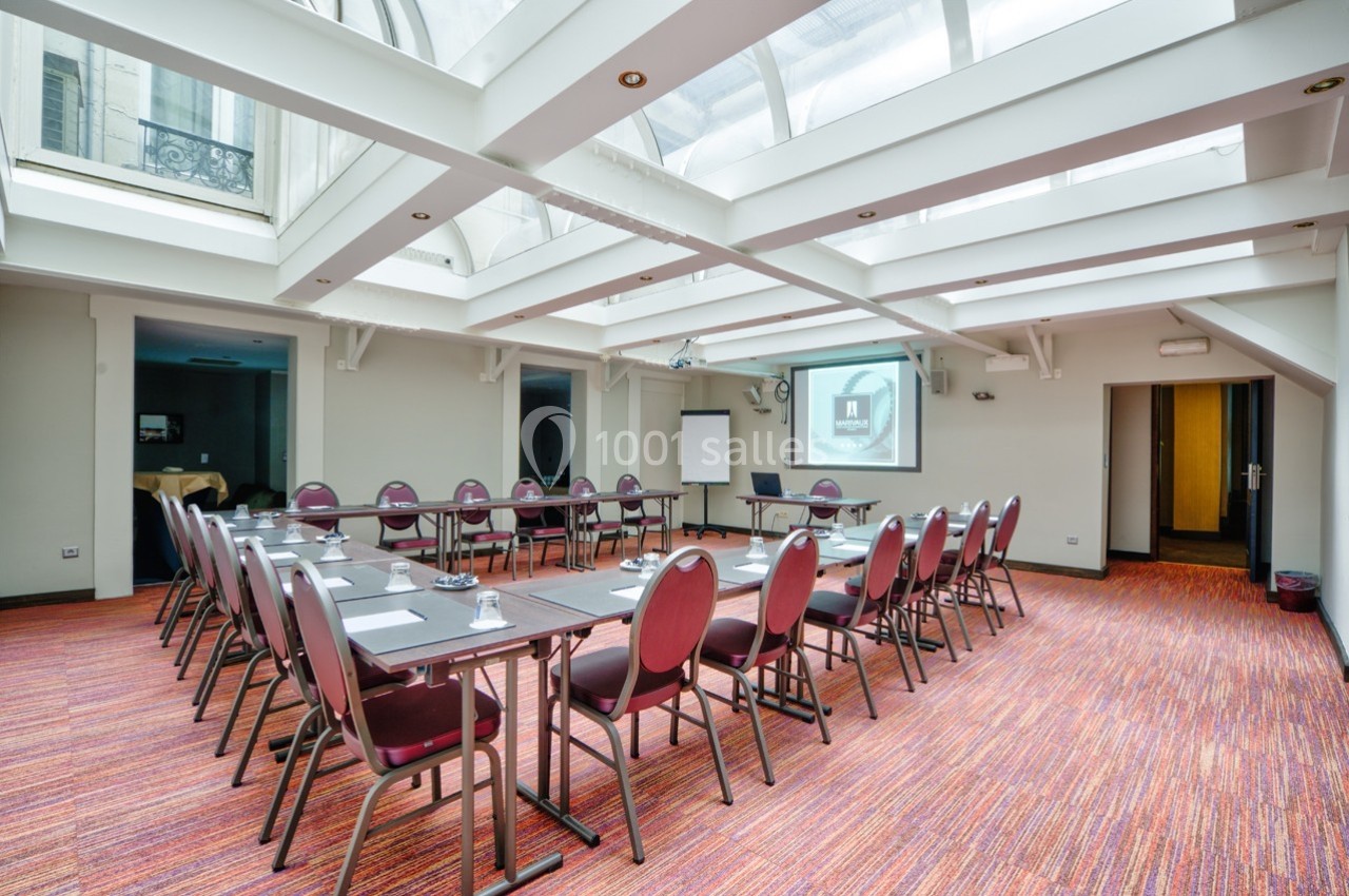 Salle de réunion lumineuse avec tables disposées en U, chaises rouges, projecteur et tableau blanc.