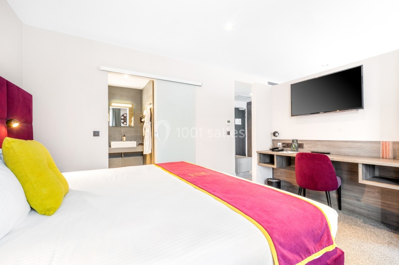 Chambre d'hôtel lumineuse avec lit double, bureau, chaise rouge, télévision murale et salle de bain attenante.