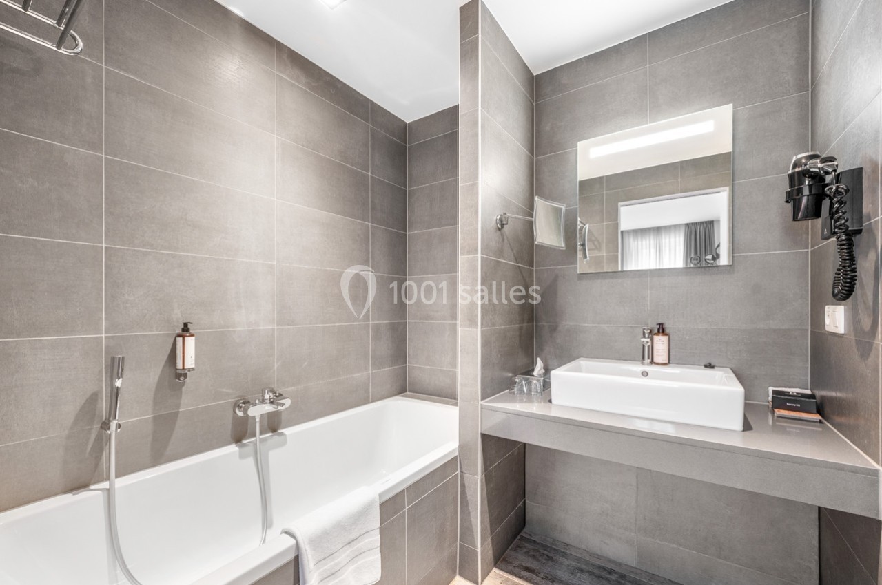 Salle de bain moderne avec baignoire, lavabo, miroir éclairé et murs en carrelage gris.