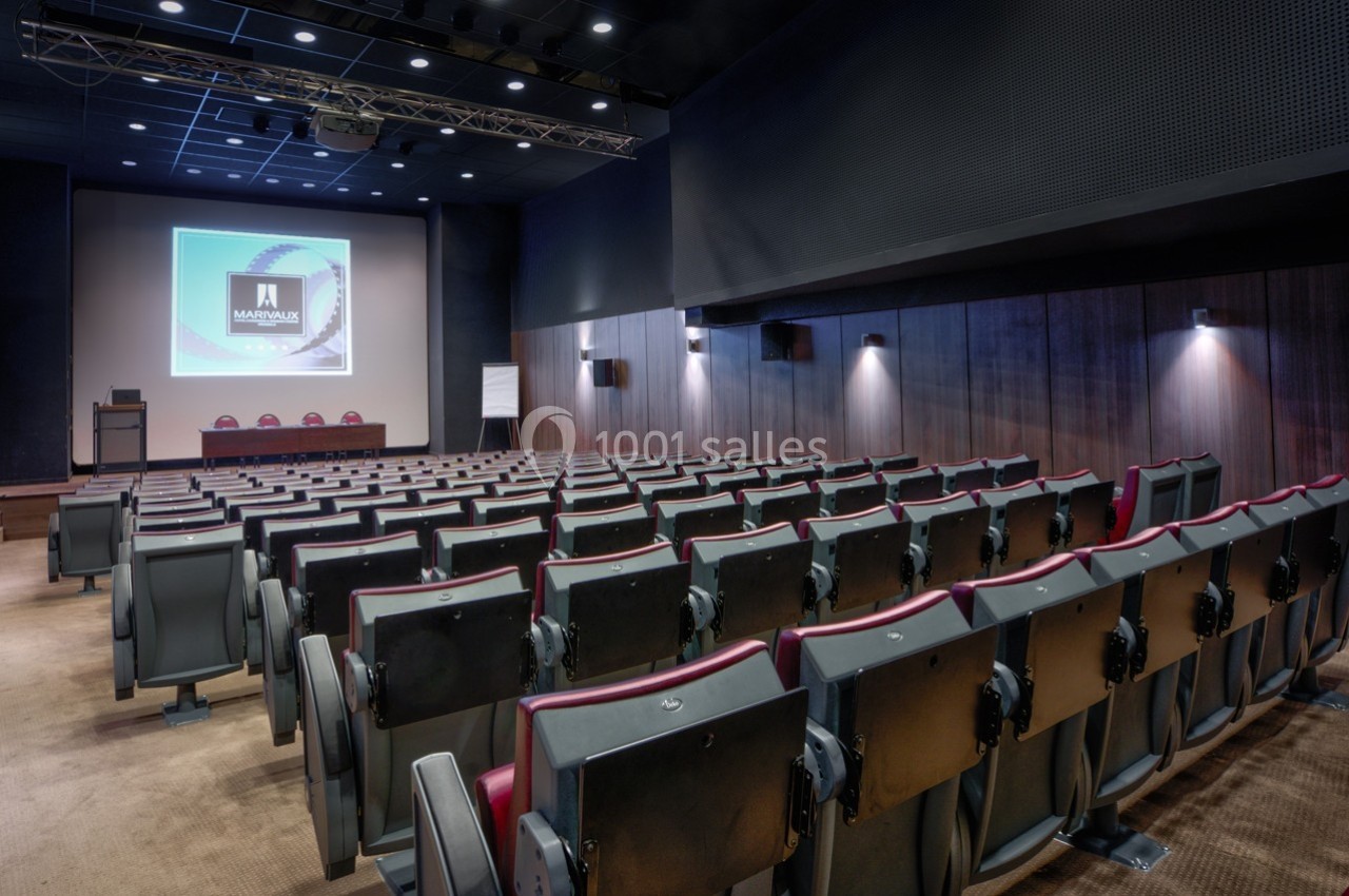Salle de conférence vide avec rangées de sièges, écran de projection allumé et éclairage tamisé.