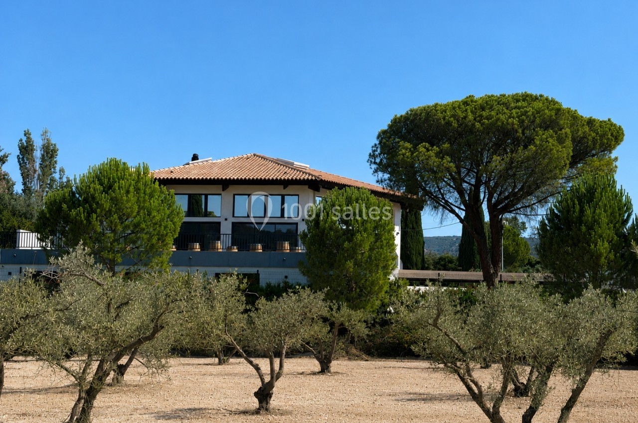 Maison moderne avec toit en tuiles, entourée d'arbres et d'oliviers, sous un ciel bleu clair.