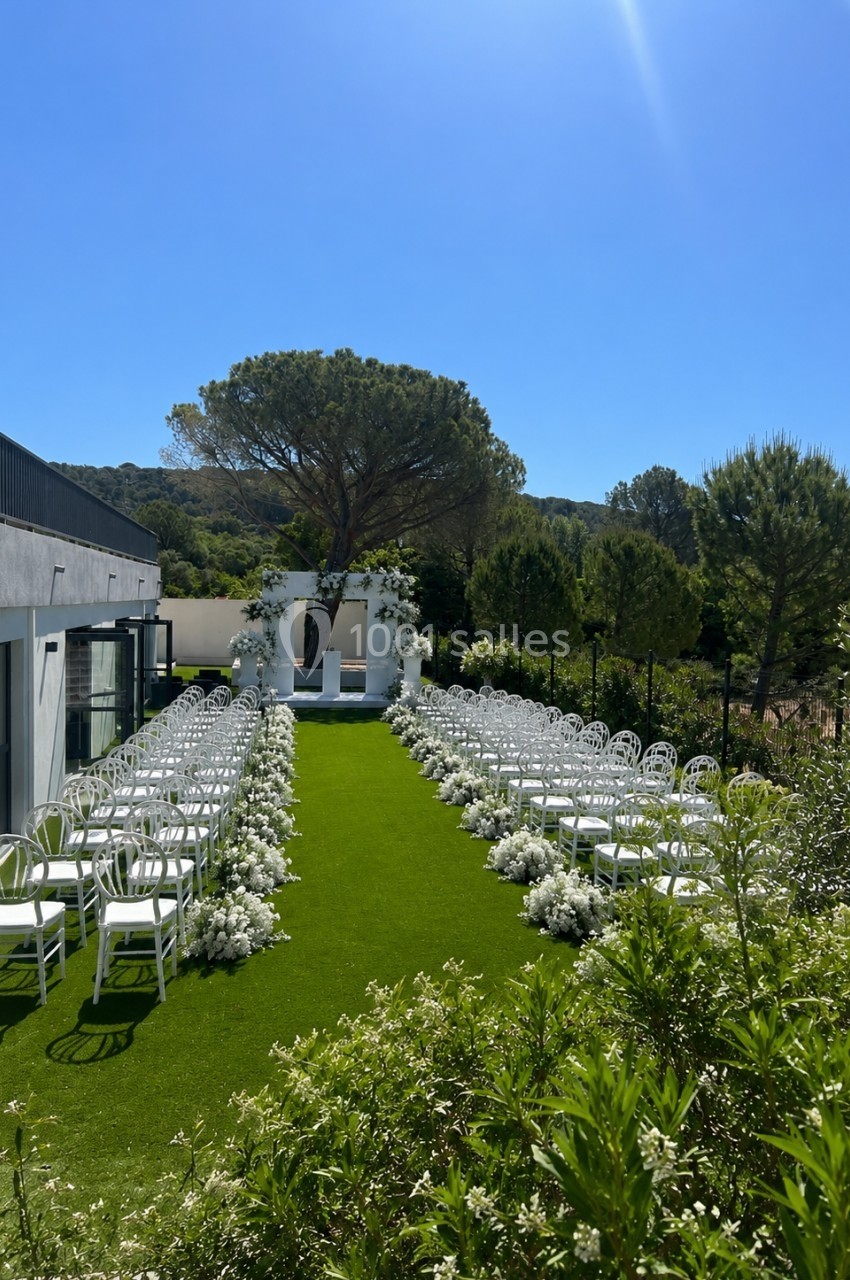 Allée centrale décorée de fleurs blanches menant à une arche de mariage sur une pelouse, entourée de verdure.