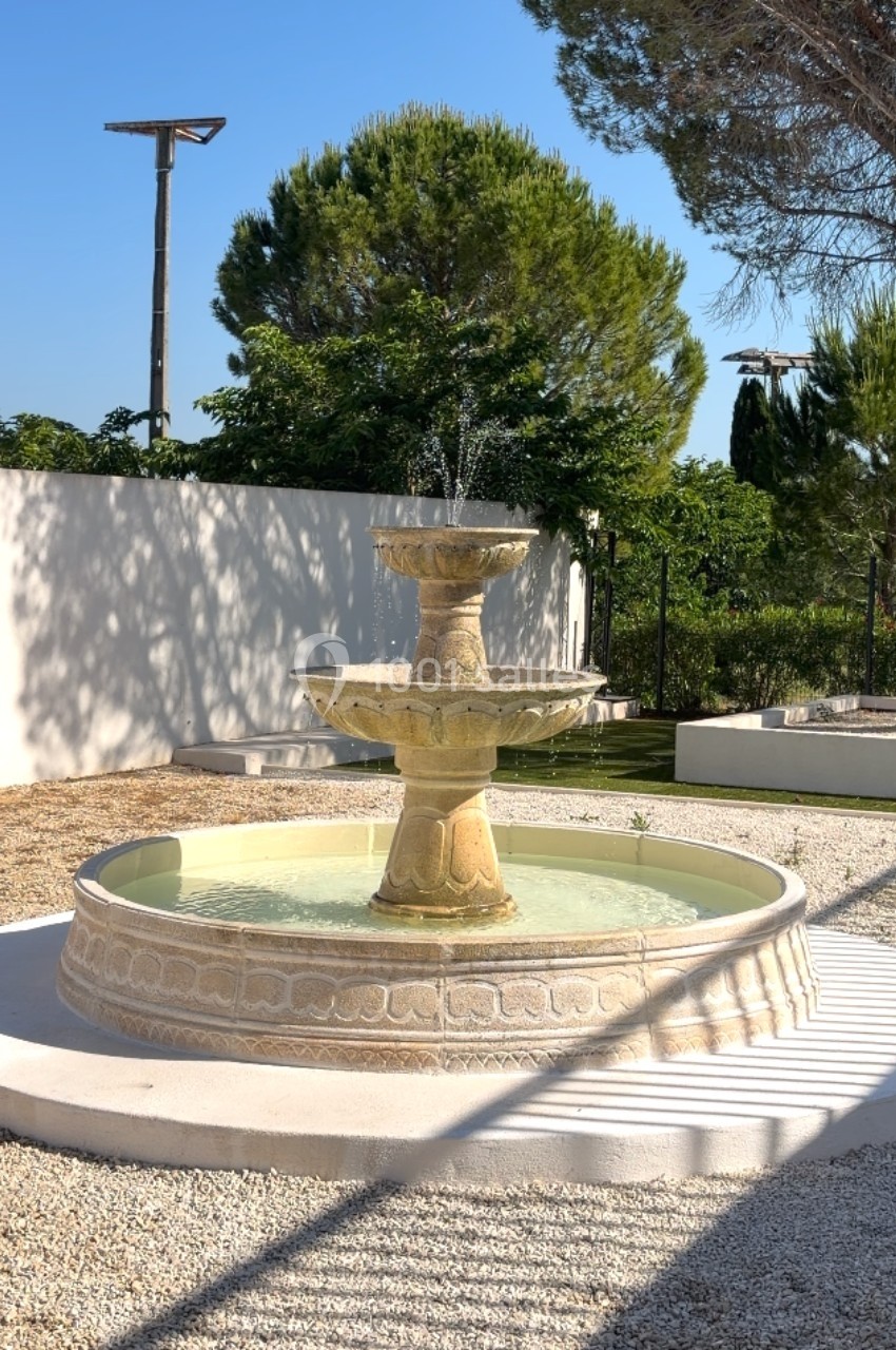 Fontaine en pierre à trois niveaux entourée de gravier, située dans un jardin avec des arbres en arrière-plan.