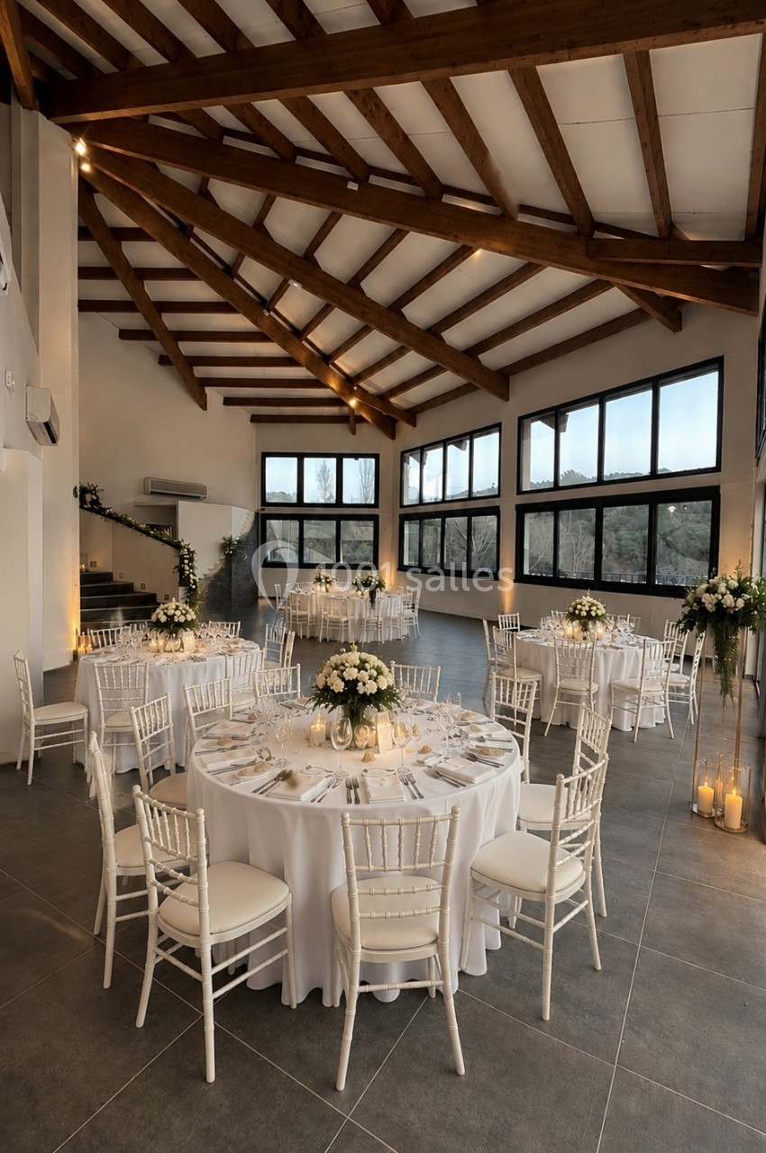 Salle de réception lumineuse avec tables rondes dressées, chaises blanches et grandes baies vitrées donnant sur un paysage.