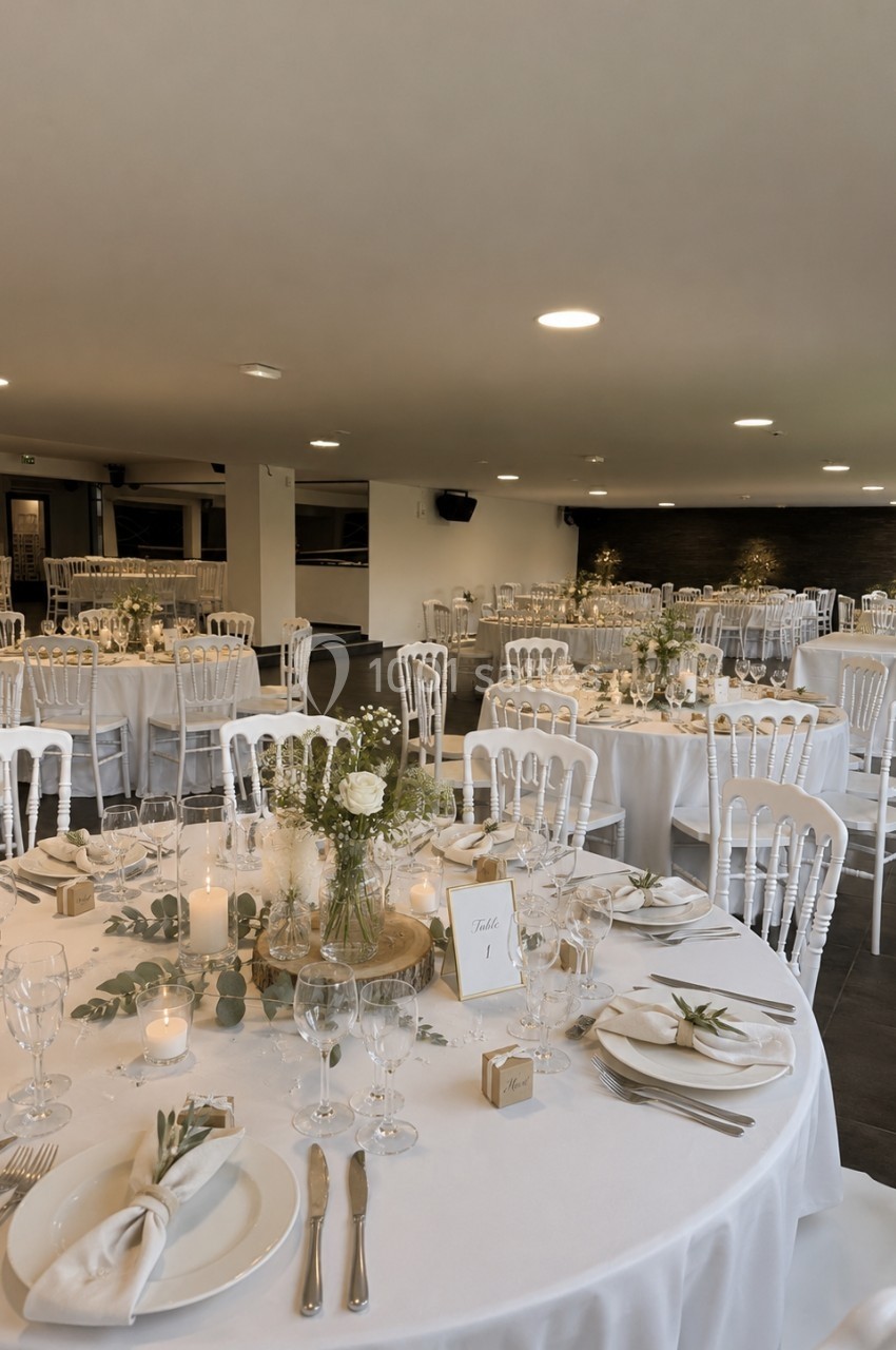 Salle de réception décorée pour un mariage, avec tables rondes dressées, nappes blanches et compositions florales.