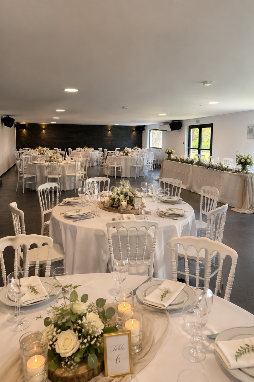 Salle de réception décorée avec des tables rondes, nappes blanches, centres de table floraux et chaises blanches.