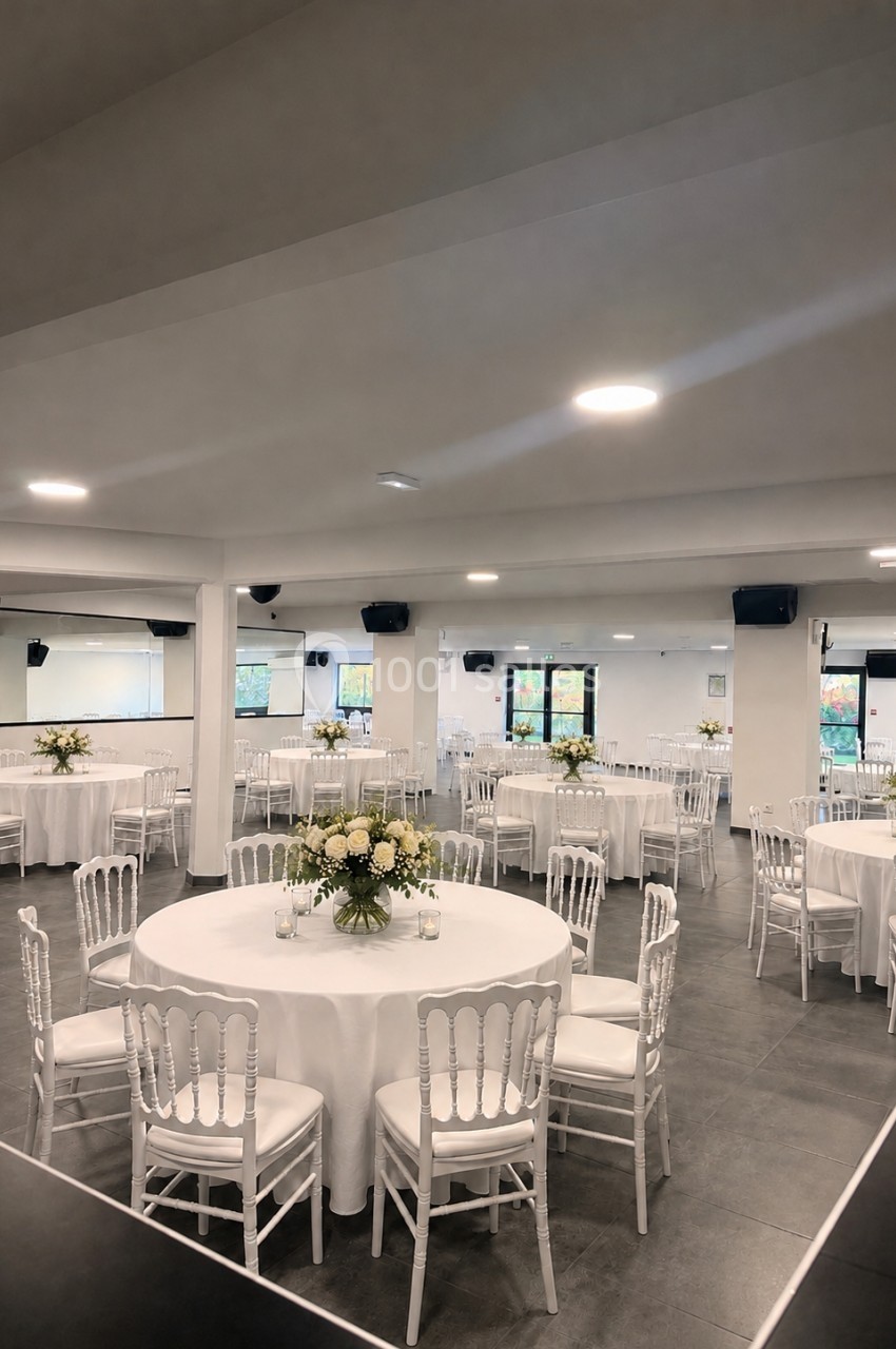 Salle de réception lumineuse avec tables rondes dressées de nappes blanches et décorées de bouquets de fleurs.