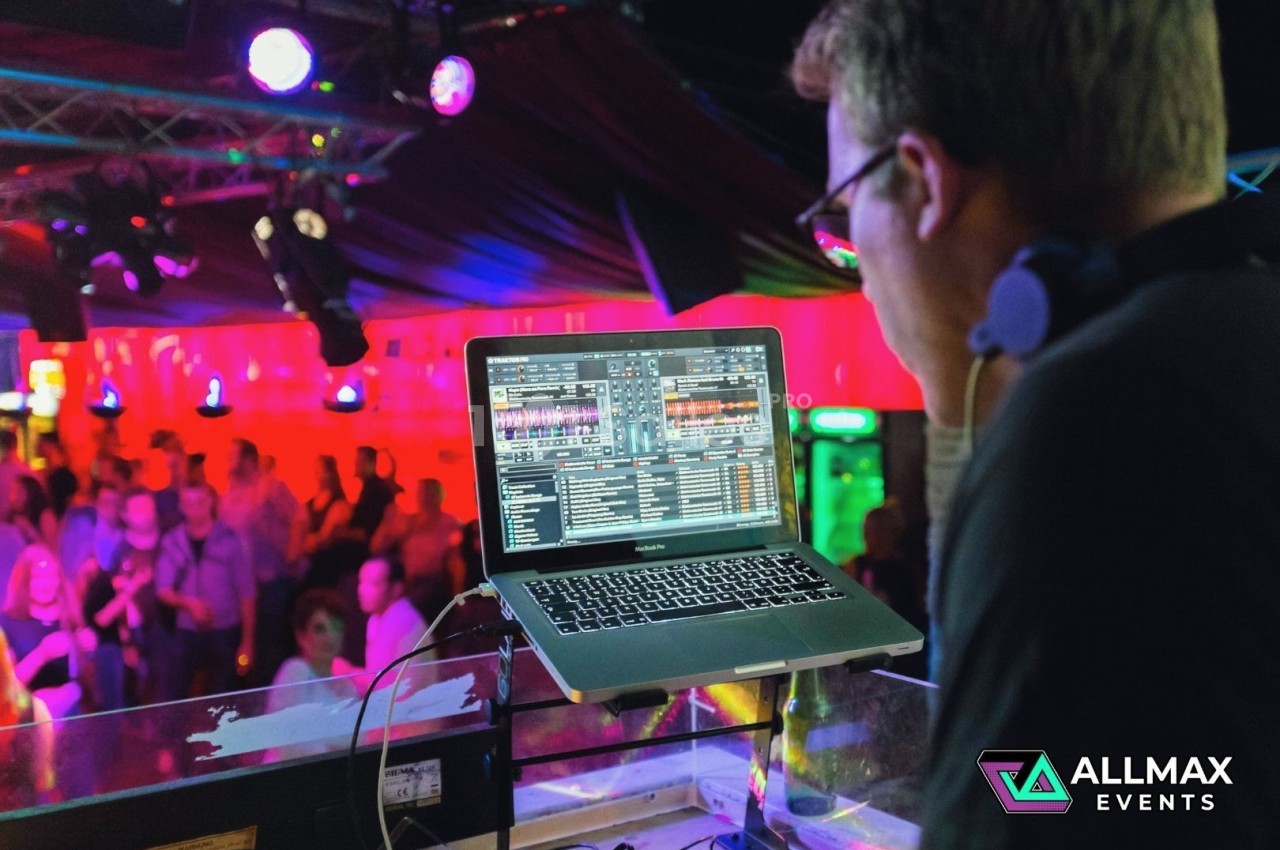 Un DJ mixe de la musique sur un ordinateur portable dans une salle éclairée par des lumières colorées, avec un public en…