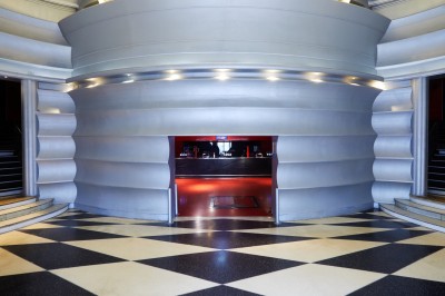 Hall d'entrée avec murs aux formes ondulées, sol à motifs géométriques et escalier menant à une porte noire.