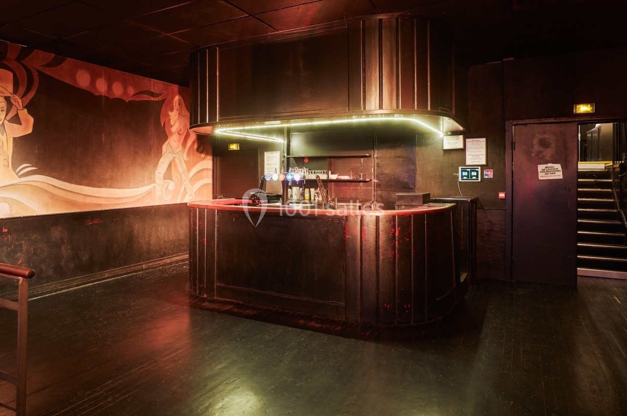 Bar en bois sombre avec éclairage tamisé, décor mural artistique et escalier visible à droite.