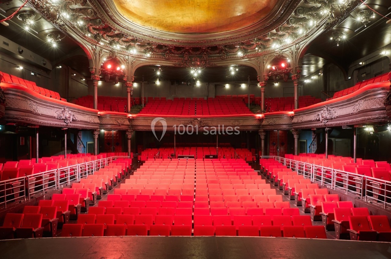 Salle de théâtre vide avec des sièges rouges, un balcon orné et un plafond décoratif éclairé.