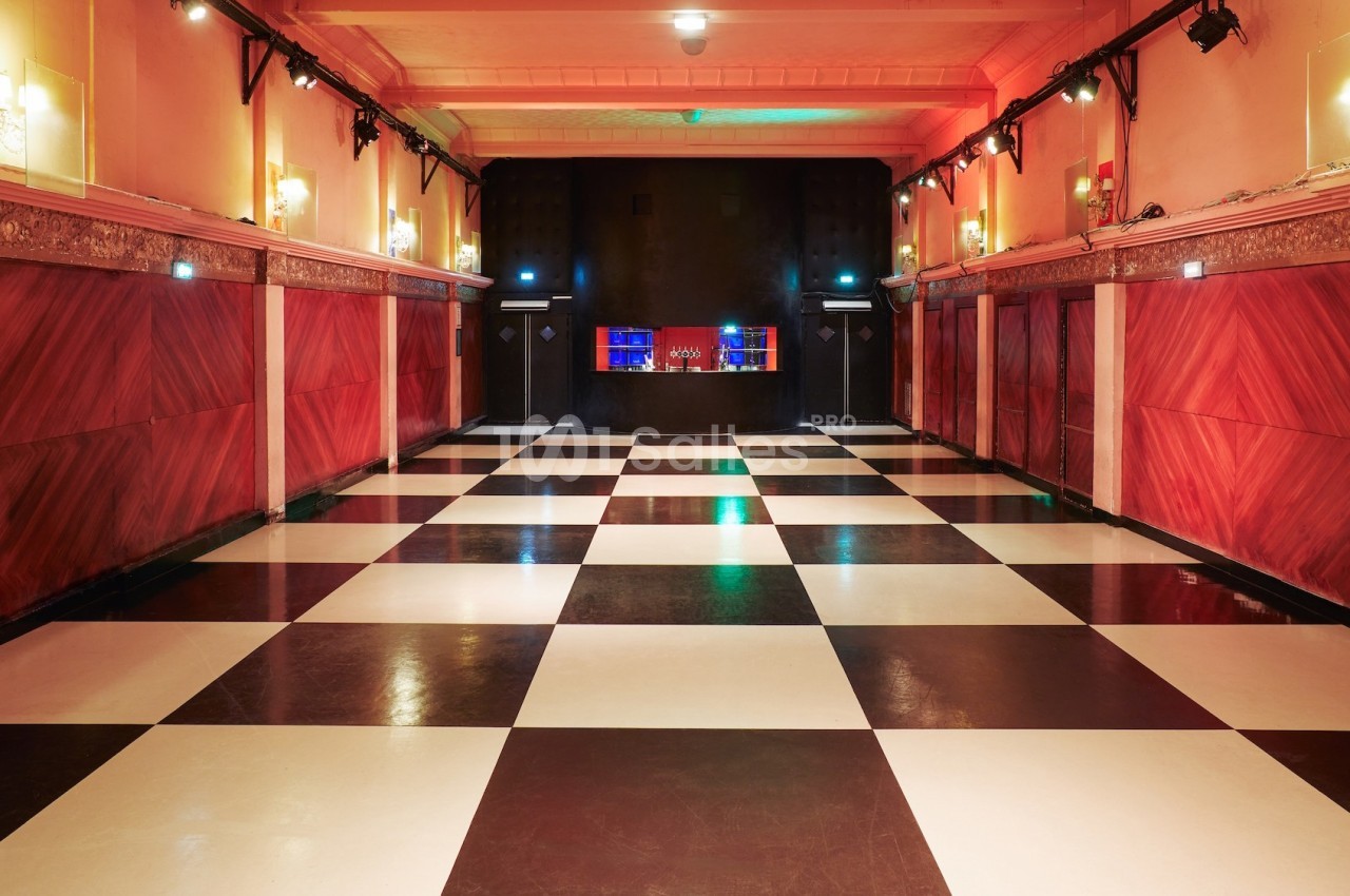 Salle vide avec sol à damier noir et blanc, murs rouges ornés de moulures et bar éclairé en arrière-plan.