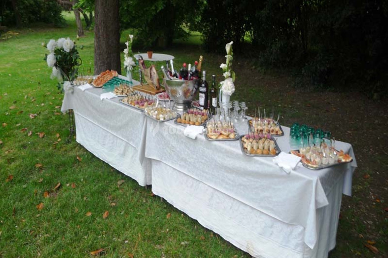 Table dressée en extérieur avec nappes blanches, proposant des amuse-bouches, boissons et décorations florales.