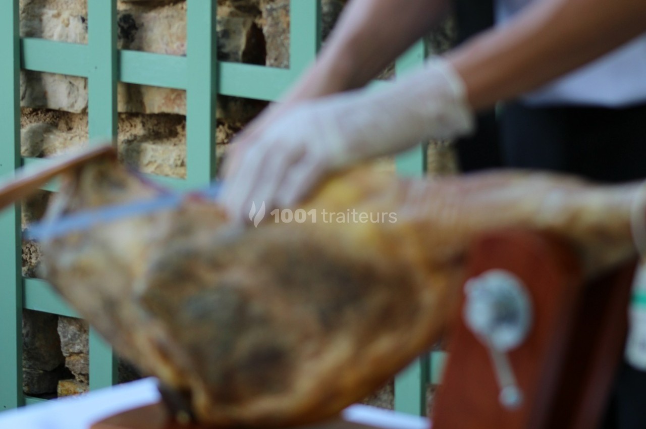 Personne découpant un jambon sec sur un support en bois, portant des gants pour une manipulation hygiénique.