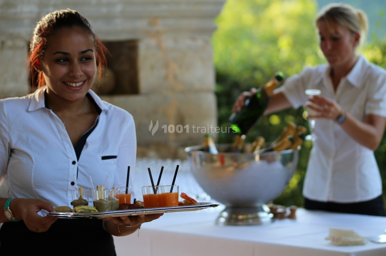 Une serveuse souriante porte un plateau de boissons, tandis qu'une autre personne sert du champagne en arrière-plan.