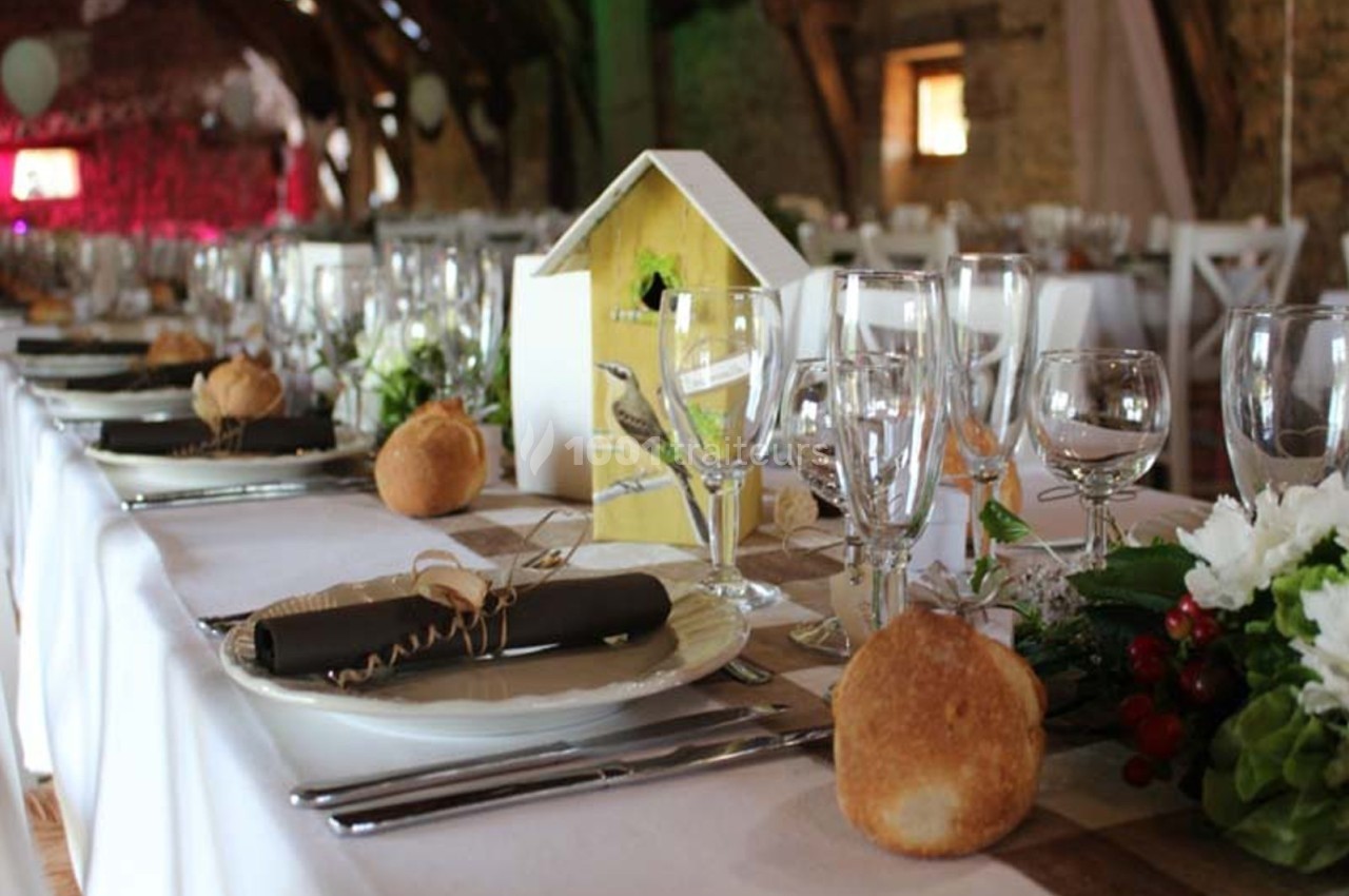 Table dressée pour un repas festif dans une salle rustique, avec vaisselle élégante, décorations et pain individuel.