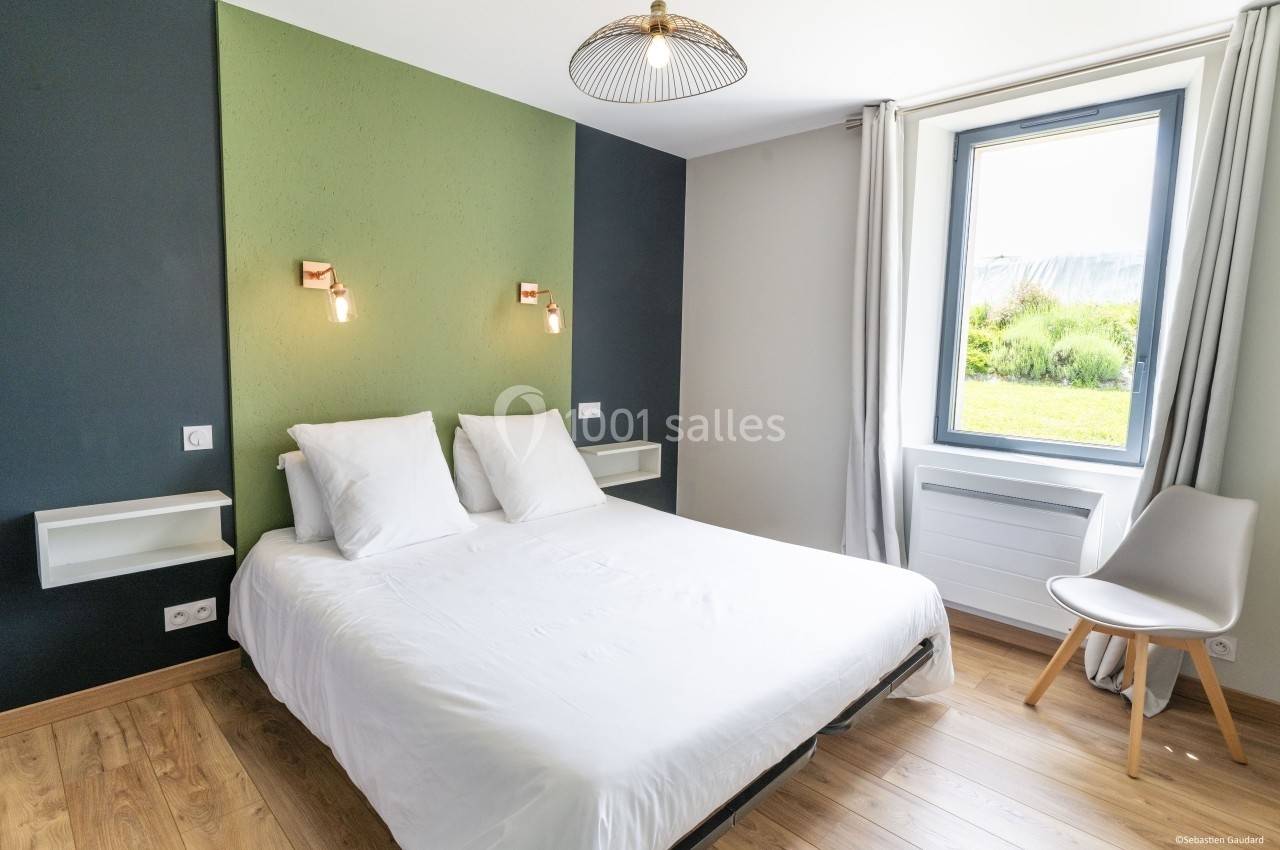 Chambre lumineuse avec lit double, mur bicolore vert et bleu, fenêtre donnant sur un espace extérieur verdoyant.