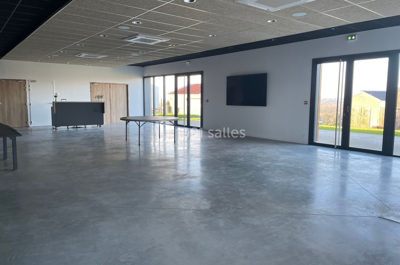 Salle vide avec sol en béton, grandes baies vitrées, écran mural et mobilier minimaliste.