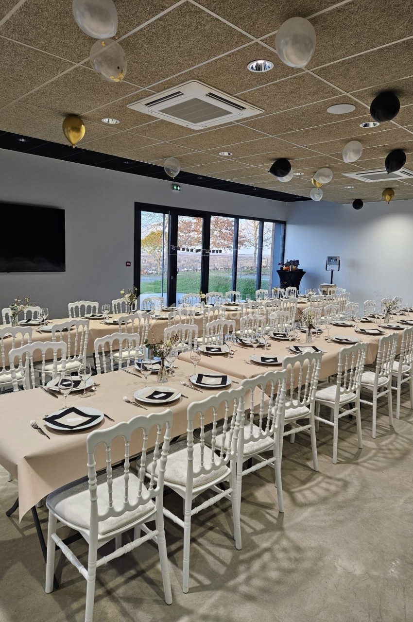 Salle de réception décorée avec des tables alignées, nappes beiges, chaises blanches et ballons suspendus au plafond.