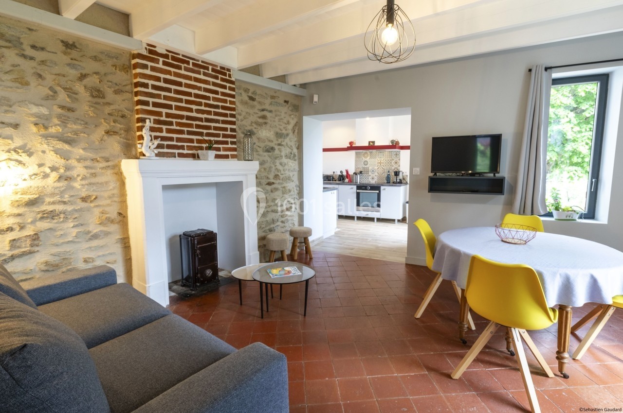 Salon lumineux avec murs en pierre, cheminée, canapé, table ronde avec chaises jaunes et vue sur une cuisine ouverte.