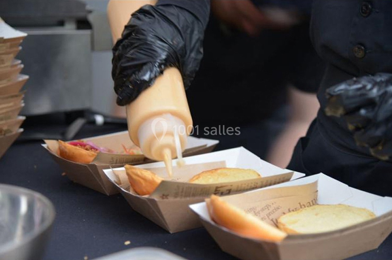 Une personne verse une sauce dans des barquettes contenant des pains à burger, sur un plan de travail.