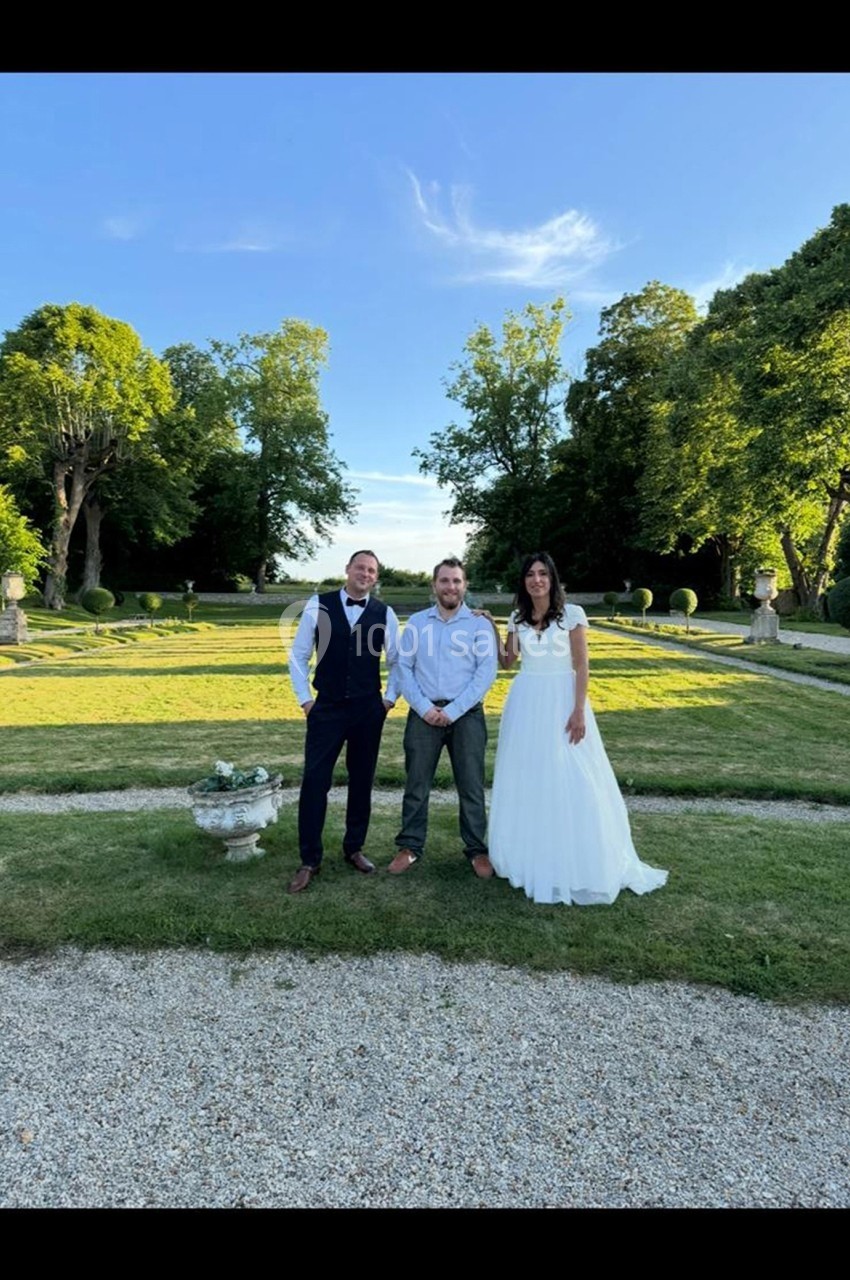 Un homme en costume, un homme en chemise et une femme en robe blanche posent dans un jardin verdoyant.