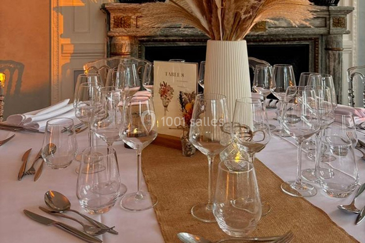 Table élégamment dressée avec nappes blanches, verres, couverts et vase contenant des herbes séchées.