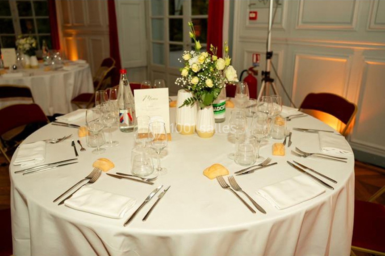 Table ronde dressée pour un repas, avec nappes blanches, couverts, verres, petits pains et centre de table floral.