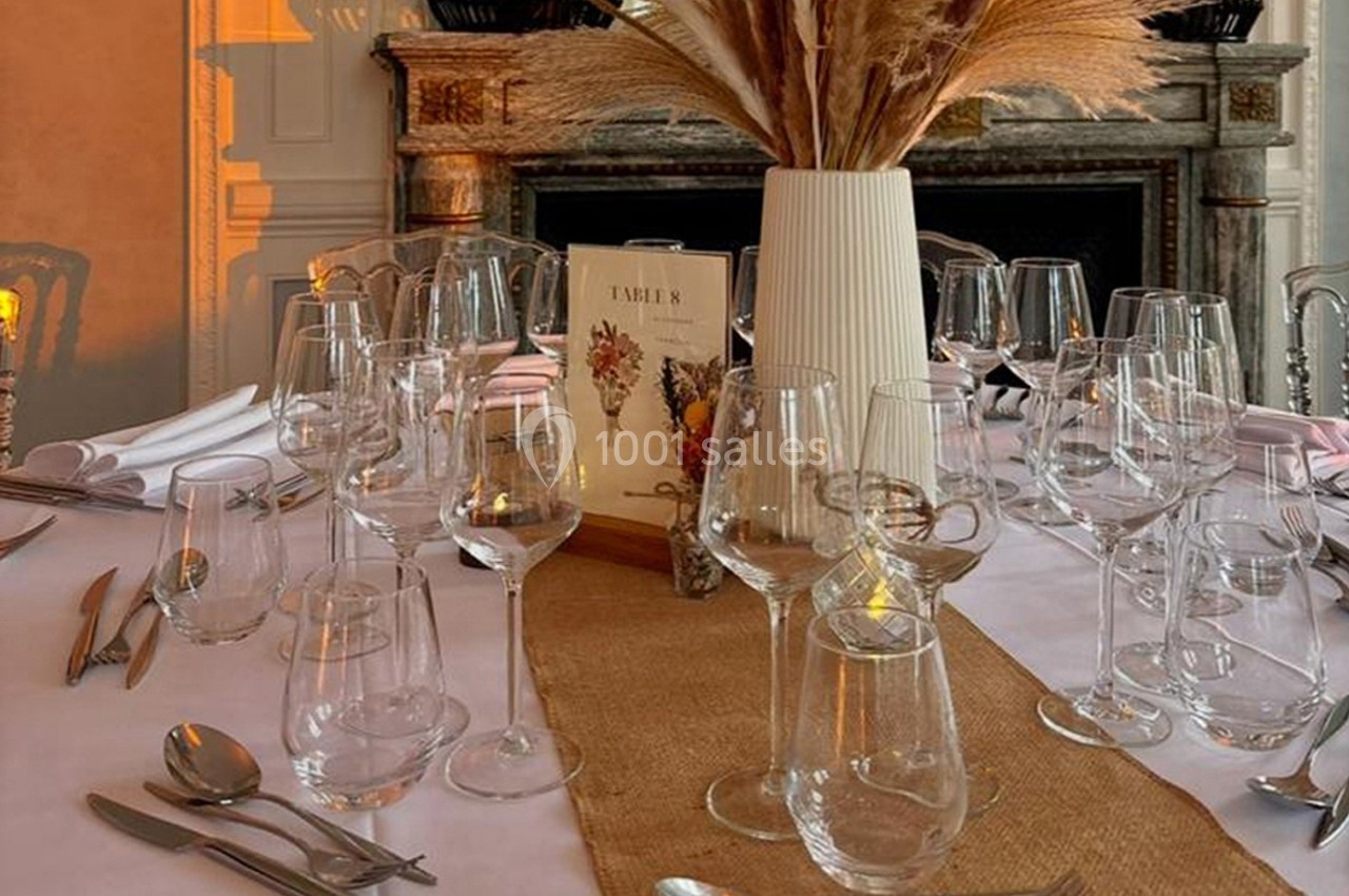 Table élégamment dressée avec des verres, couverts, un chemin de table en toile de jute et un vase de fleurs séchées.