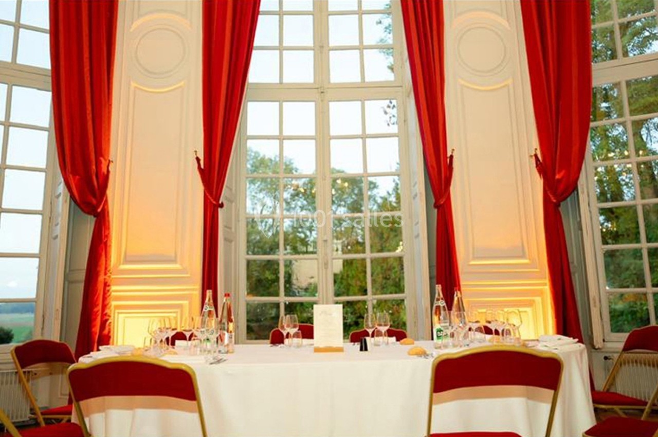 Table dressée avec nappes blanches, chaises rouges et grandes fenêtres encadrées de rideaux rouges dans une salle lumineuse.