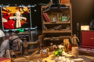Miniature Shooting photo / Tournage Atelier de création avec une table remplie de matériaux, une caméra suspendue et une poupée en tissu en cours de fabrication.