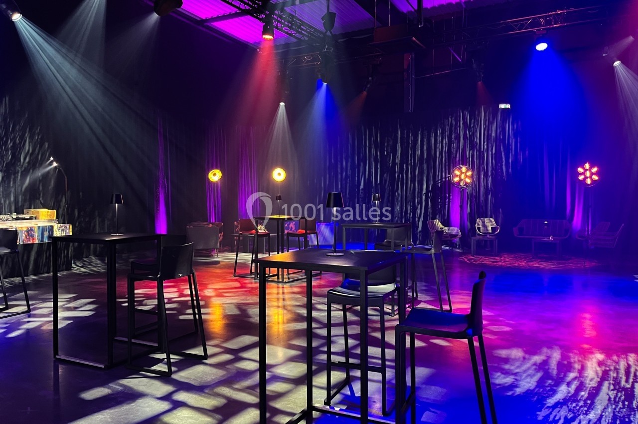 Salle sombre aménagée avec des tables hautes, des chaises et des éclairages colorés créant une ambiance festive.