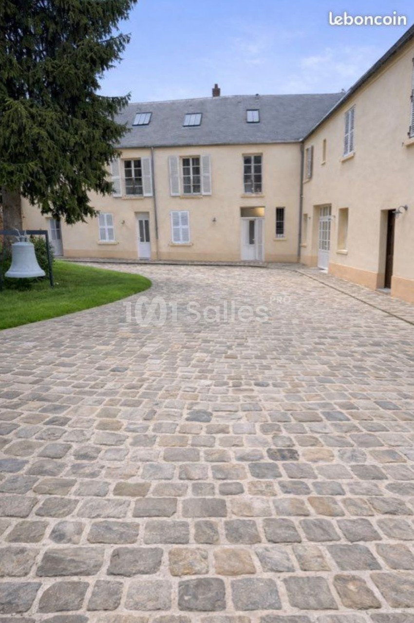 Cour pavée en pierre entourée de bâtiments bas aux façades crépies beige clair, volets blancs et toiture en ardoise, avec une cloche blanche sur la pelouse à gauche. Let me verify length: roughly 165 characters, too long. Need ≤125. Reformulate: "Cour