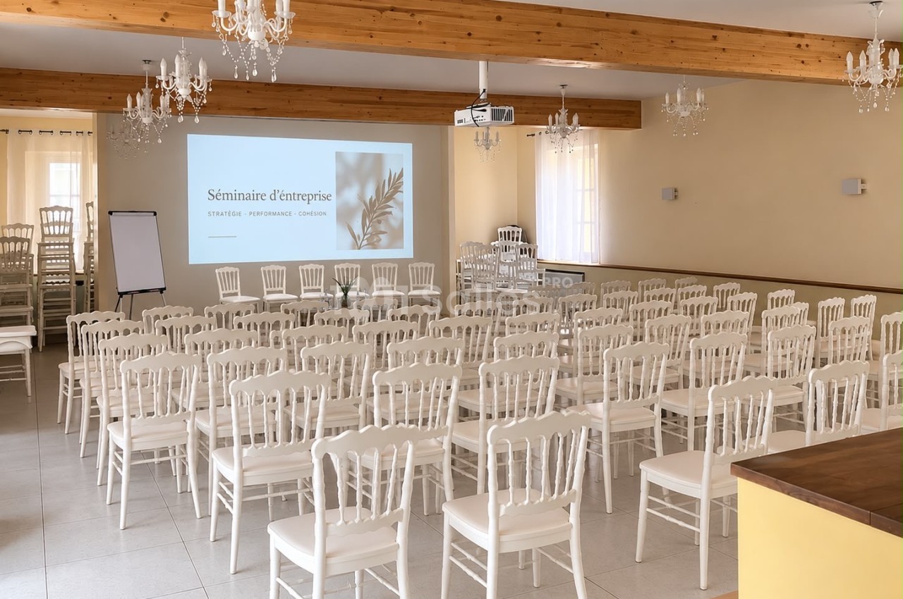 Salle dressée en conférence, rangées de chaises blanches face à un écran de projection sous des poutres apparentes.