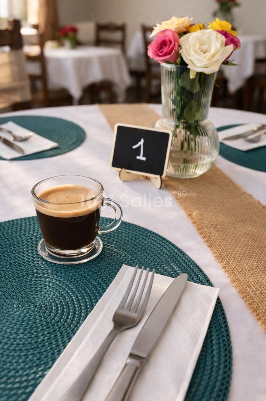 Table ronde dressée avec set vert, couvert, tasse de café en verre, vase de roses et numéro de table 1 sur chemin de jute.