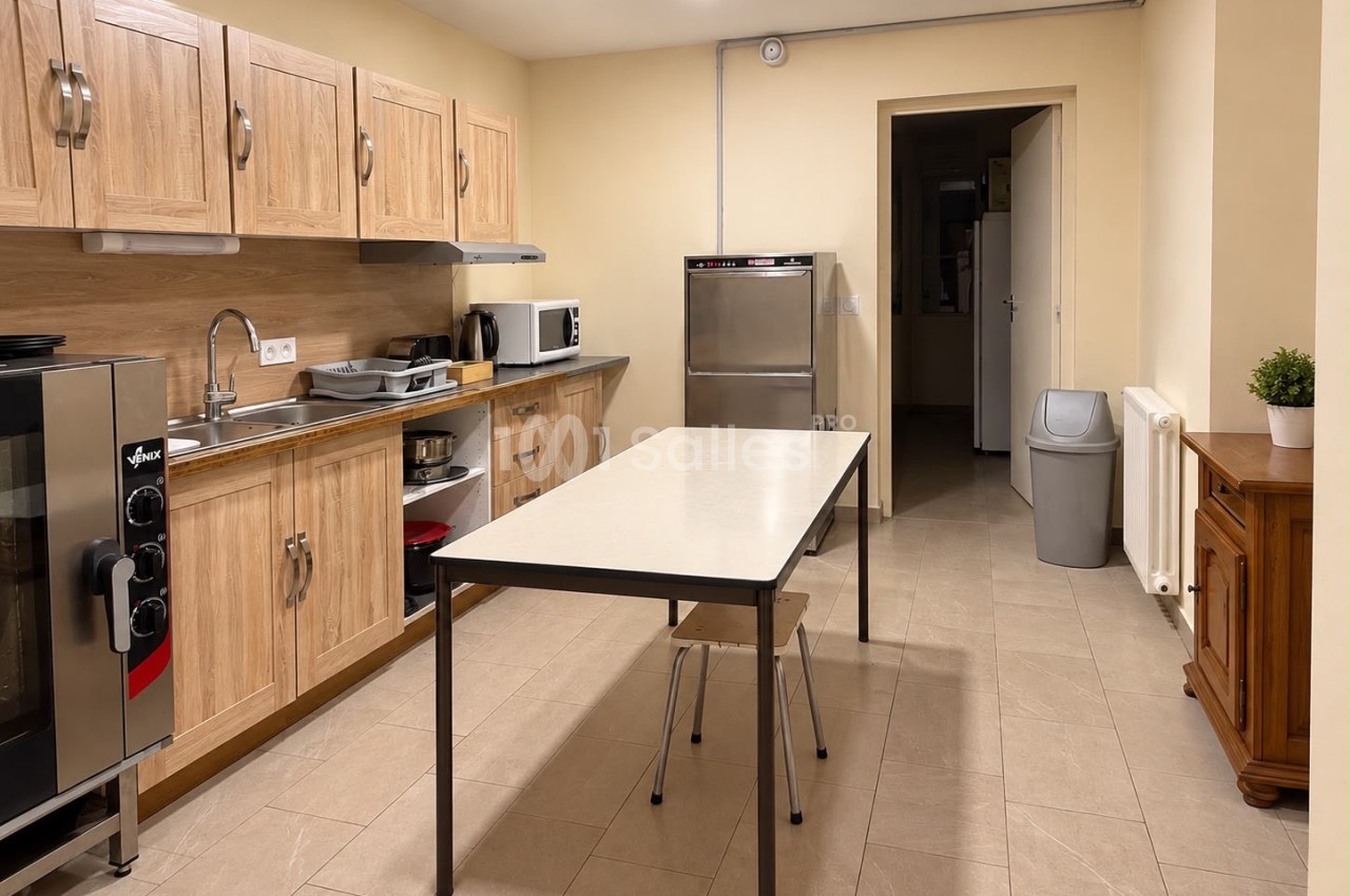 Cuisine professionnelle aménagée avec plan de travail en bois clair, four mixte inox à gauche, lave-vaisselle inox, et une table rectangulaire au centre.