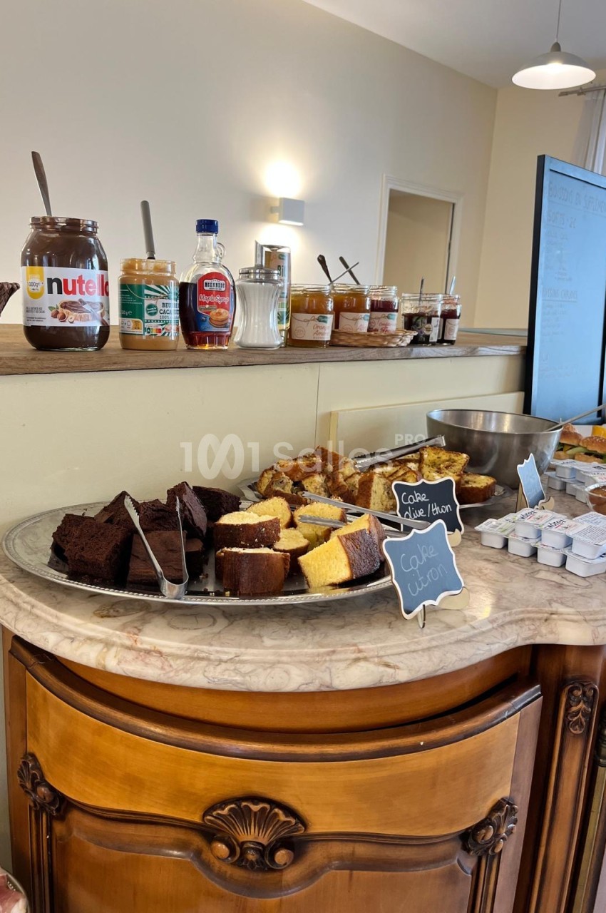 Buffet sucré sur un comptoir en marbre : brownies, tranches de cake, pâte à tartiner au chocolat et pots de confiture.