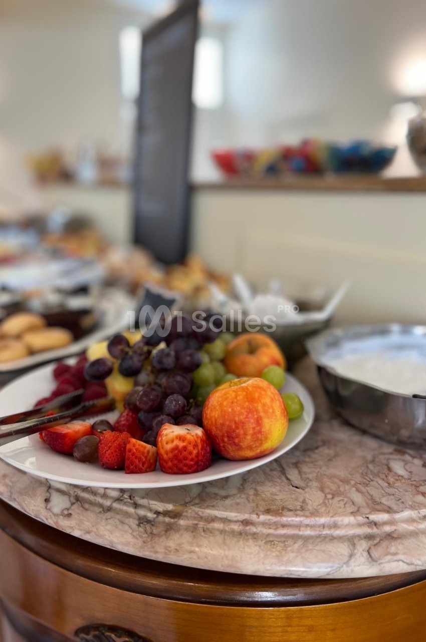Assiette de fruits frais (raisins noirs et verts, fraises, pêches, pomme) posée sur une table en marbre, sur fond de buffet flou.