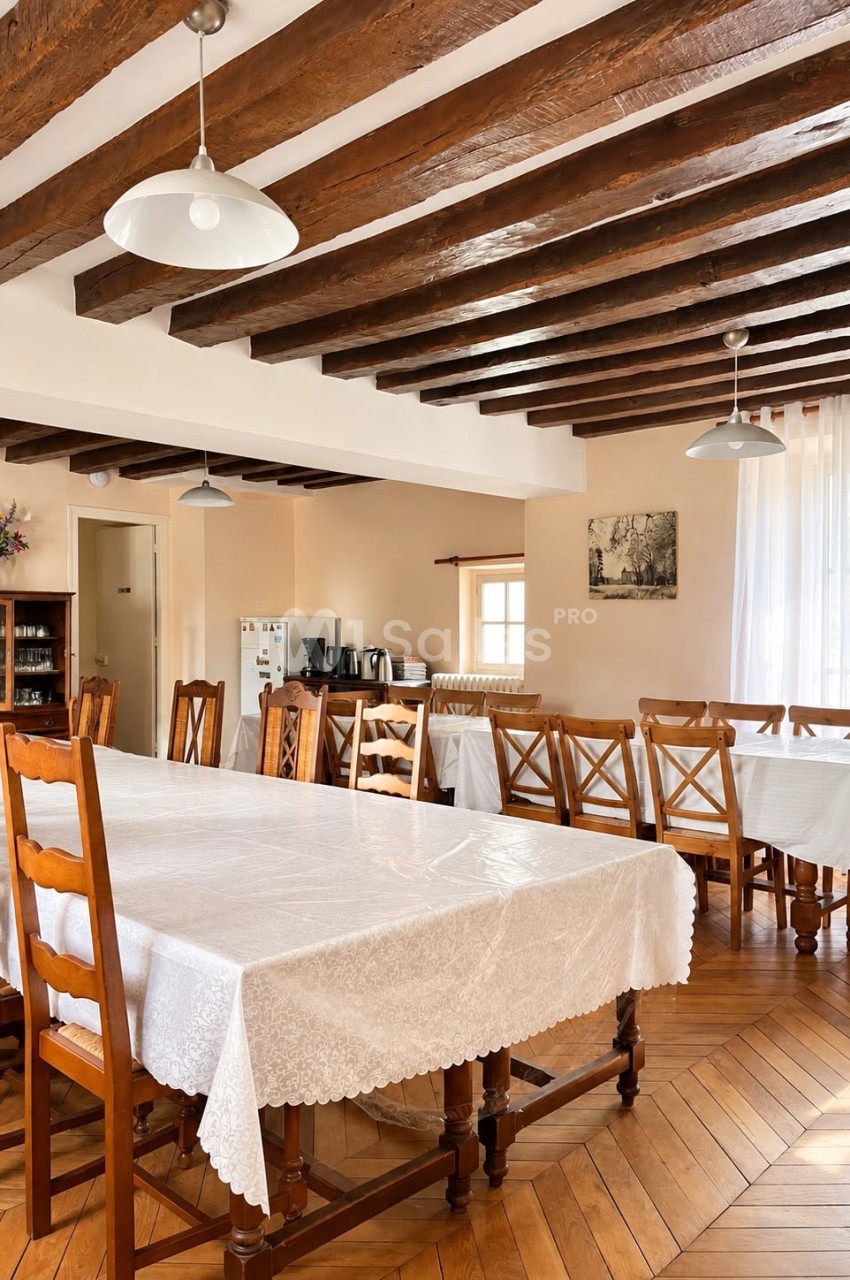 Salle à manger rustique avec poutres apparentes en bois, plusieurs grandes tables recouvertes de nappes blanches entourées de chaises en bois, et parquet en chevrons.